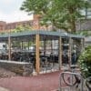 20 40 Security Cycle Enclosure bike storage area with numerous bicycles parked inside, surrounded by a metal fence. The structure is located in an urban setting with a building and trees visible in the background. Additional bike racks are situated outside the storage area.