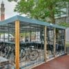 40 space security cycle enclosure bicycle parking area with a metal fence encloses several parked bicycles. Trees and a building with large windows are visible in the background, along with a tall clock tower. The area is paved with a dark surface.