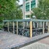 A bike storage area filled with bicycles is surrounded by a metal fence with a gray roof and wooden support posts. The storage is adjacent to a brick pathway and located near a multi-story building with large windows and surrounded by trees.