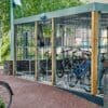 A secure bike shelter with a metal fence and gate houses multiple bicycles. The shelter, supported by wooden beams, has a gray roof and sits on a red pathway in a leafy outdoor area. A lone bike is parked outside the shelter.