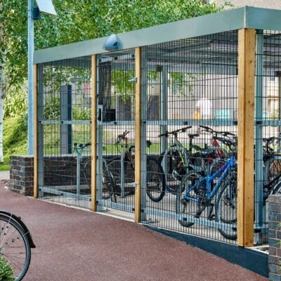 A secure bike shelter with a metal fence and gate houses multiple bicycles. The shelter, supported by wooden beams, has a gray roof and sits on a red pathway in a leafy outdoor area. A lone bike is parked outside the shelter.