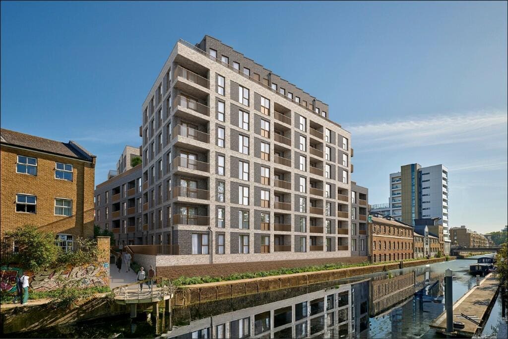 A modern multi-story residential building with a brick facade is situated adjacent to a canal. The structure features numerous balconies and large windows. Other buildings, some old and some new, are visible nearby, and a clear blue sky is overhead.