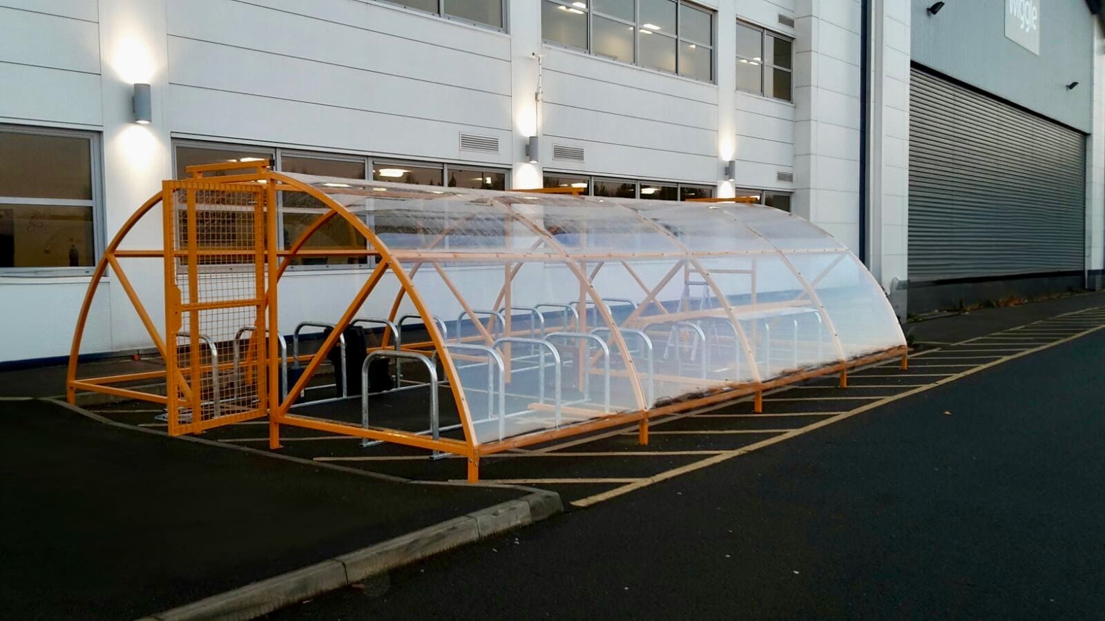 A transparent and orange bike shelter with metal benches located in front of a modern industrial building. The shelter is well-lit, with parking space markings visible on the dark pavement.