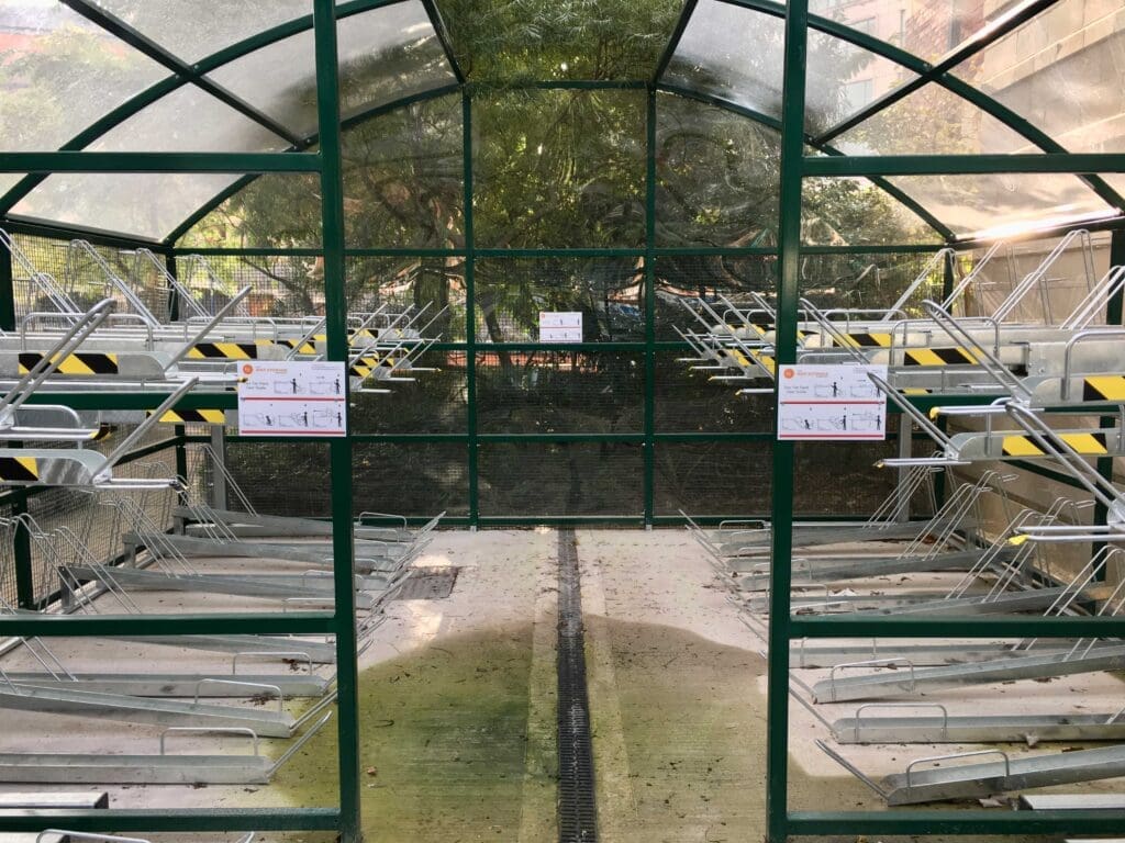 A metal-framed structure with transparent walls and roof, presumably a greenhouse or storage area. Inside, numerous metal racks are arranged along both sides, with some signs posted on them. The ground is covered in concrete and the back shows a green background.