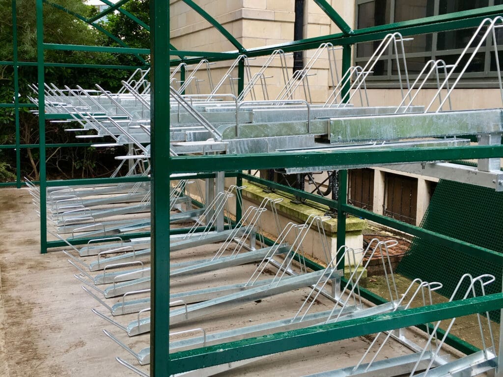 Empty metal bicycle racks are arranged in two rows on a concrete surface. The racks are installed under a green, metal-framed shelter near a building. The area is clean and organized, indicating it is ready for use.