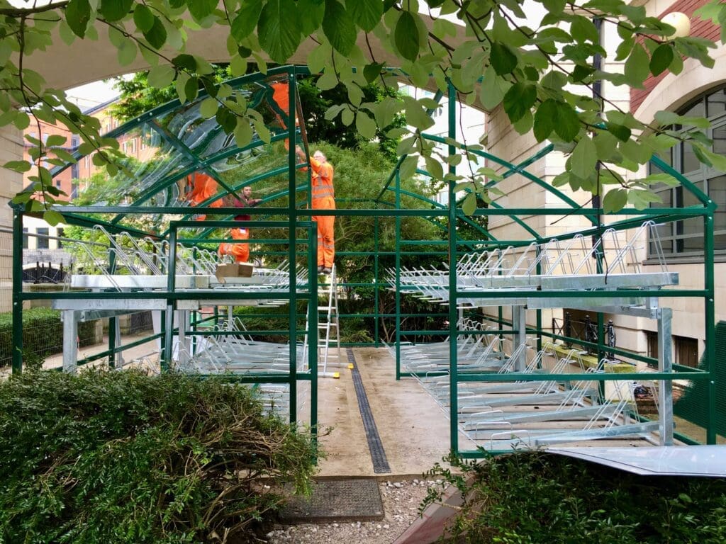 A partially constructed bike storage frame standing under an archway. Green metal structures are visible with no glass panels attached. There is some greenery around and inside the frame, along with construction materials and a ladder placed centrally.