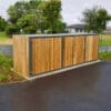 A wooden cycle locker with vertical slats stands on a paved area near a green space and a street. The enclosure, featuring timber options, houses an Amazon Eco 2 Cycle Locker. Surrounding the secure bike storage are trees, some grass, and a few buildings in the background.
