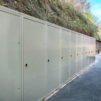 Rows of grey vertical bike lockers are aligned next to a pathway. The background features a steep hillside covered with greenery.