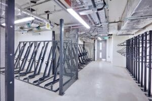 Brightly lit indoor bike storage facility with multiple metal bike racks lining the walls on both sides. The ceiling features exposed pipes and electrical wiring. The room is spacious, clean, and organized, with an exit door visible at the far end.