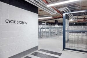 A white brick wall with a sign on it, with a cycle storage facility in the background with a mix of two tier racks and stands.