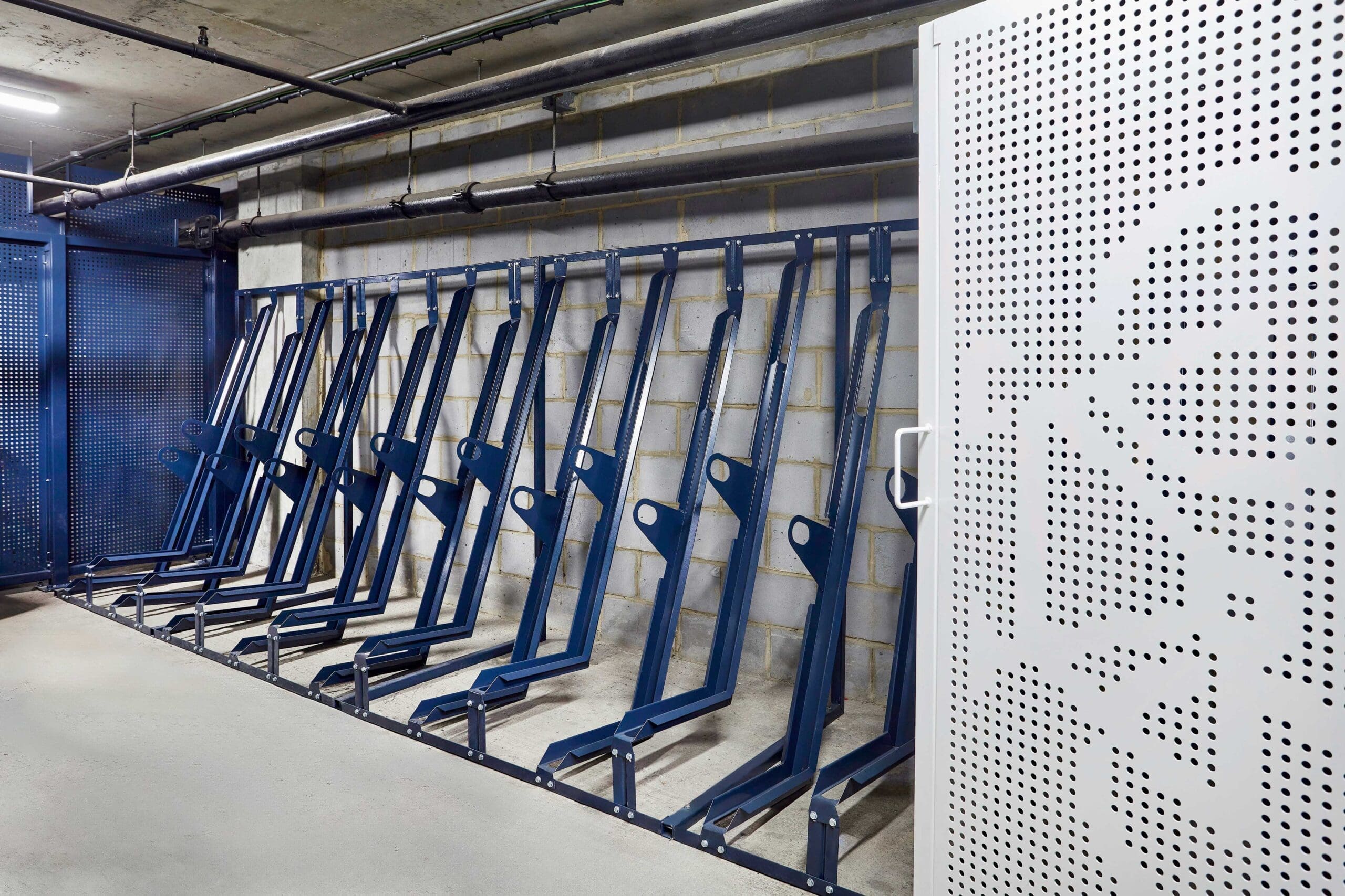 An indoor bike storage area with multiple blue semi vertical bike racks aligned against a concrete wall. The space is well-lit, with a perforated metal door featuring abstract patterns visible on the right. Pipes and structural elements are seen overhead and on the walls.
