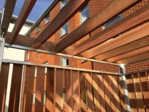 A wooden bin store structure with an open slatted roof is under construction next to a modern brick building with multiple windows. Sunlight casts shadows on the wood, giving a pattern effect. The sky is clear and blue in the background.
