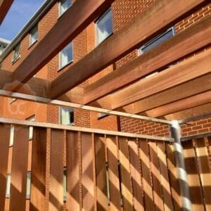 Wooden pergola with horizontal beams overhead and vertical slats on the side, situated near a brick building with multiple windows. The sky is clear and blue, casting shadows from the pergola structure onto the surrounding area.