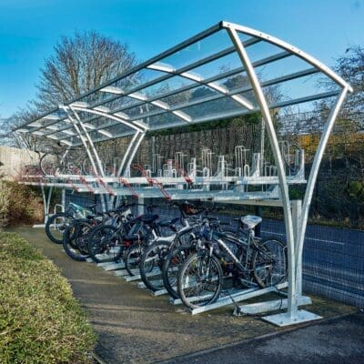 A modern outdoor Chelsea Bike Shelter with a transparent roof features a double-decker bike rack system. Several bicycles of different styles and colors are parked in the lower and upper racks. The area is surrounded by greenery and bathed in sunlight.