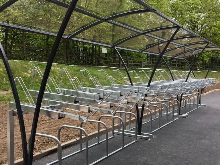 A covered bike rack with multiple individual bike slots, located next to a pathway and surrounded by greenery. The rack features a transparent roof supported by metal arches, offering protection from the elements.