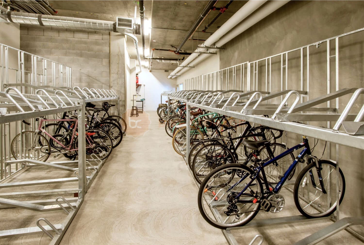 A bike storage facility with multiple bicycles parked on both sides of a long, narrow corridor. The bicycles are secured in tiered metal racks, and the space is well-lit with concrete walls and ceiling, and visible pipes running along the ceiling.