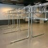 A row of empty, metal bike racks in a modern, indoor setting with grey flooring and white walls. The racks are lined up neatly in parallel, with multiple hooks but no coats or other items hanging from them. The room appears clean and well-lit.