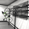 A green road bike is hanging vertically by its front wheel on an industrial-style bike rack attached to a white brick wall. The bike rack is made of black metal and includes several curved hooks for multiple bicycles. The flooring is gray and the setting appears to be a garage or storage area.