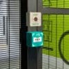 A close-up of a security gate with a "DOOR RELEASE" button and a green emergency exit button. The gate features black metal bars and a partially visible bright green wall in the background.