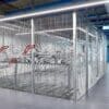 A large, well-lit room containing several rows of metal two tier bike racks enclosed by the anti-climb mesh fencing. The ceiling features exposed pipes and beams.