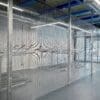 The image shows a secured indoor bicycle storage area with anti-clim mesh fencing. Inside, empty bike racks are arranged along the walls under overhead fluorescent lighting. The flooring is smooth and grey, and there are visible metal pipes and structural beams on the ceiling.