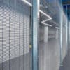 A secure indoor area with a security mesh fence and sturdy steel beams, featuring an empty corridor with polished concrete floors and a ceiling with linear LED lighting. The atmosphere appears industrial and modern.