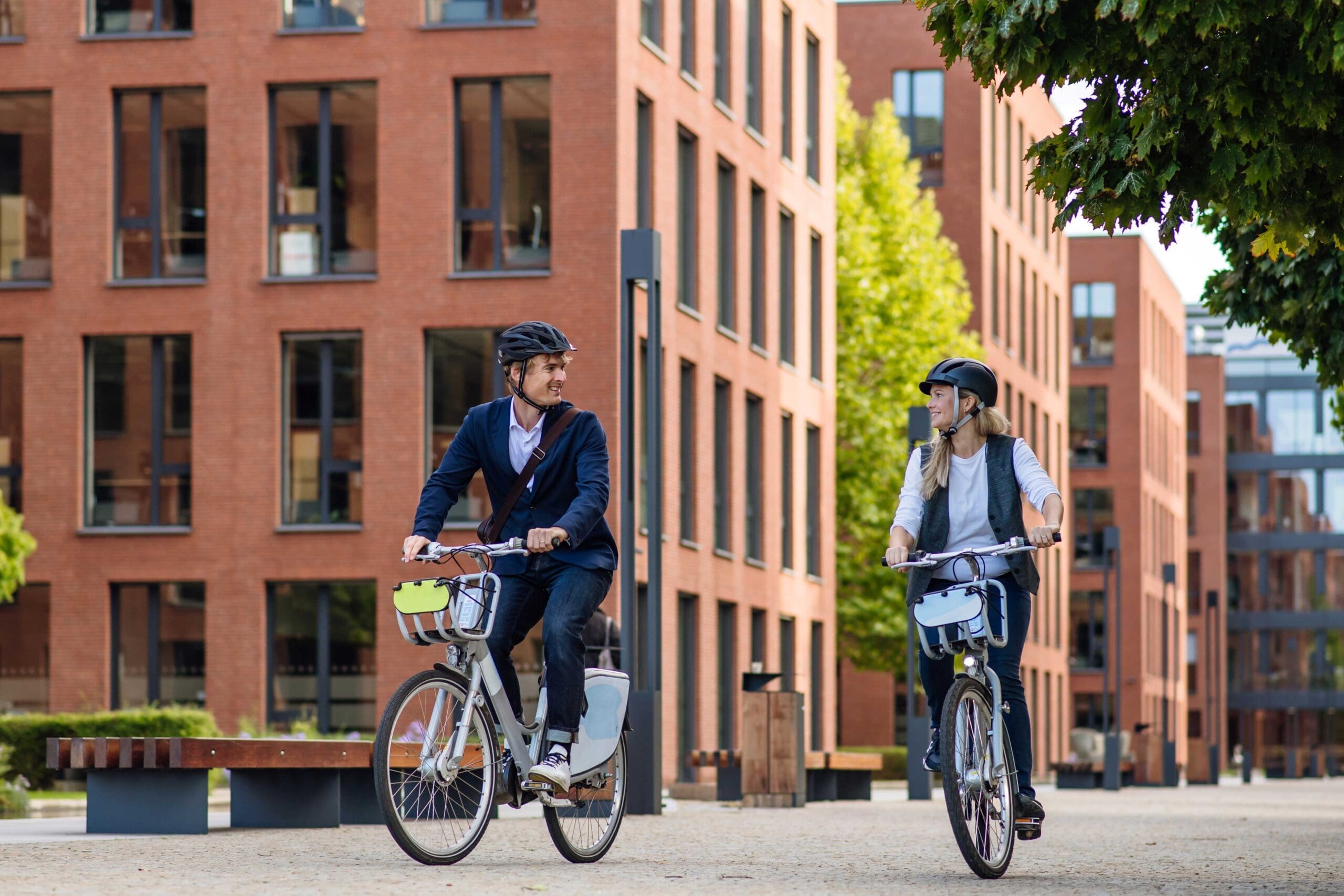 Two people are riding bicycles in an urban area with tall red-brick buildings. They are both wearing helmets and casual clothing, looking at each other and smiling. The scene is well-lit with a tree and a bench visible in the background.