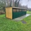 A long outdoor wooden structure with a black roof and green lower panels, set on a concrete slab surrounded by grass. Designed for secure bike storage, it features weather-resistant materials and is located in a park-like area with trees and a cloudy sky in the background.