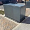 A rectangular gray metal box, featuring a three-point locking system and designed for secure bike storage, is positioned on a paved area that includes light gray and brown bricks. Several identical weather-resistant boxes are also visible in the background. The surrounding area shows a partial view of a parked car, a brick wall, and a wooden fence.