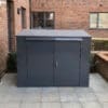 A large, dark gray metal horizontal bike locker with two front doors and a lock is placed on a tiled patio area. The box is situated against a brick building, with windows visible in the background.