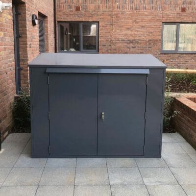 A large, dark gray metal horizontal bike locker with two front doors and a lock is placed on a tiled patio area. The box is situated against a brick building, with windows visible in the background.