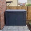 A horizontal bike locker that is positioned on a tiled area adjacent to a brick wall and wooden fence. The shed has a flat top and two front doors with a lock. It is placed in an outdoor area with some green shrubs visible in the background.