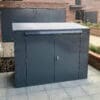 A gray metal horizontal bike locker with two front doors and a lock is placed on a tiled patio area. The box is situated against a brick building, with windows visible in the background.