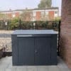 A grey horizontal bike locker that is positioned on a tiled area adjacent to a brick wall and wooden fence. The shed has a flat top and two front doors with a lock. It is placed in an outdoor area with some green shrubs visible in the background.