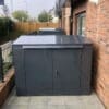 A horizontal bike locker that is positioned on a tiled area adjacent to a brick wall and wooden fence. The shed has a flat top and two front doors with a lock.