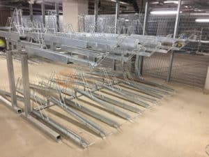 A metal bike rack with multiple tiers is installed in a concrete indoor parking area. The tiers are empty, and the structure is made of shiny, galvanized steel. The space is illuminated with overhead fluorescent lights, and a metal fence is visible in the background.