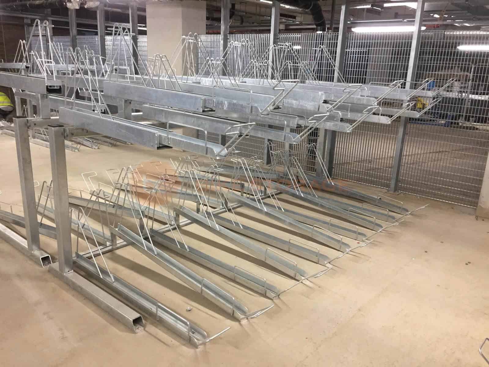 A metal bike rack with multiple tiers is installed in a concrete indoor parking area. The tiers are empty, and the structure is made of shiny, galvanized steel. The space is illuminated with overhead fluorescent lights, and a metal fence is visible in the background.