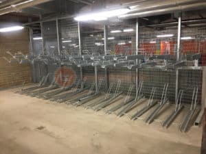 A spacious indoor bicycle storage area featuring a two-tier bike rack system. The upper and lower metal racks are empty, ready to securely hold multiple bicycles. The setting is well-lit with industrial lighting and has a concrete floor and wire mesh walls.