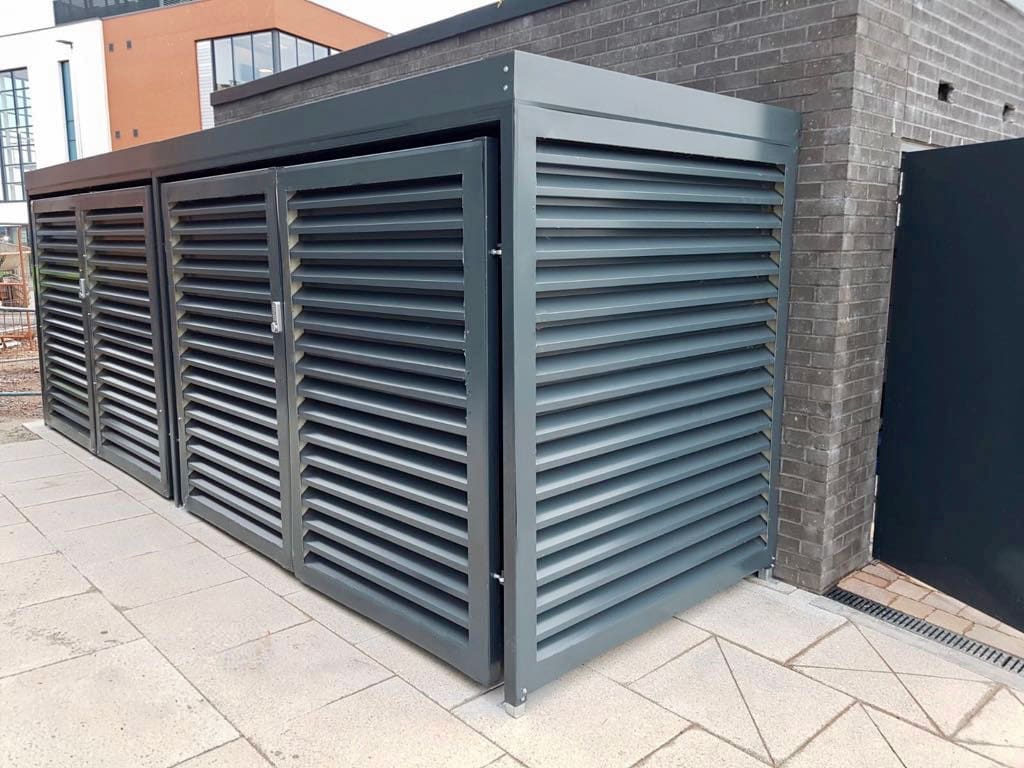 A modern metal storage unit with louvered panels stands outdoors on a tiled pathway adjacent to a brick wall. The unit features horizontal slats for ventilation and is situated in an urban setting with buildings in the background.