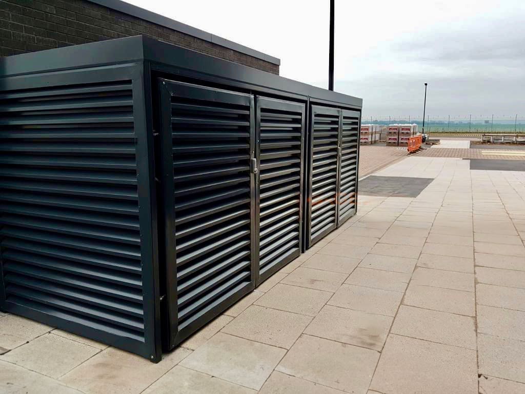 A modern, black, metal storage shed with horizontal slats stands on a tiled pavement. The shed is situated near a body of water and an orange barricade is visible in the background. The sky is overcast.