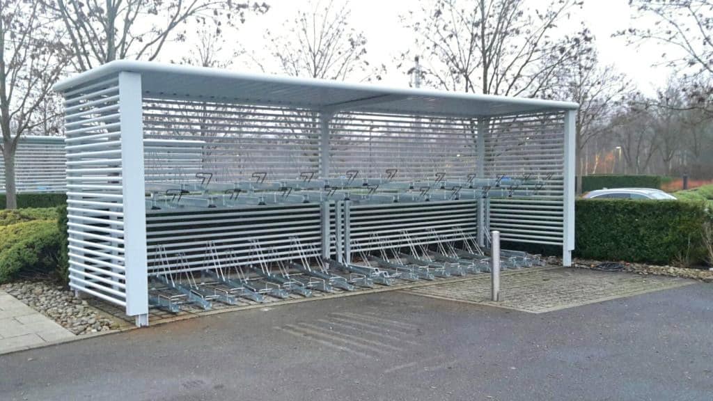 A covered bike rack with metal bars and slots for securing bicycles is situated in an outdoor area with a background of leafless trees and a paved area in front. No bicycles are currently parked in the rack.