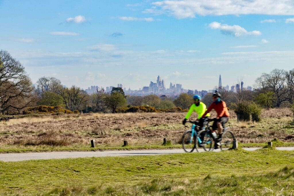 Two people riding through a green park with the London skyline in the background on a sunny day