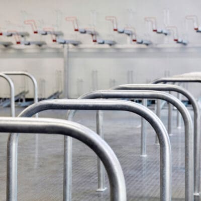 A row of empty metal bike stands inside an industrial-style room with a checkered metal floor. The bike racks are evenly spaced, and the background shows pipes and other industrial equipment. The area appears clean and well-maintained.