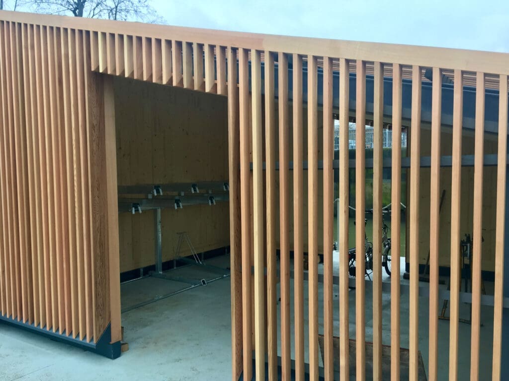 A modern wooden bike shed with vertical slats is shown. The shed has an open entrance revealing bike racks inside. The design features clean lines and a natural wood finish, situated on a concrete foundation. The background includes a partially visible overcast sky and some trees.
