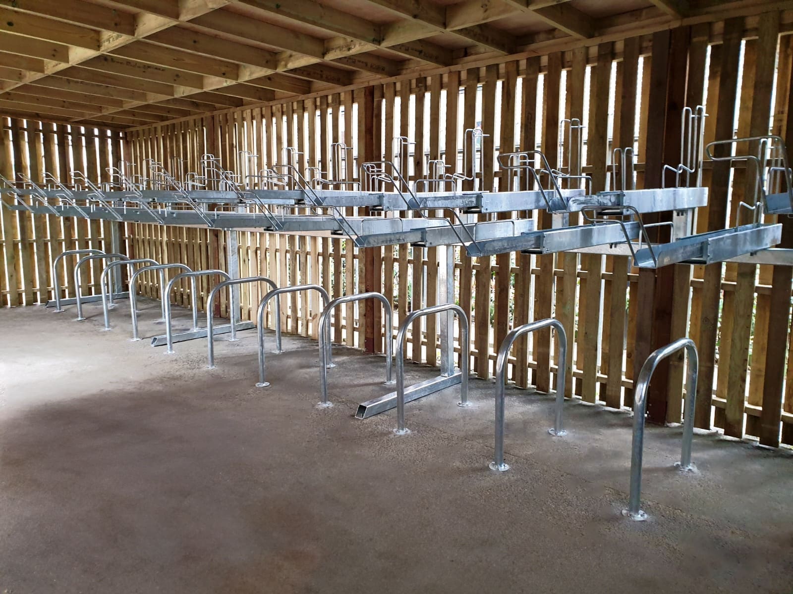 A room with wooden walls and ceiling, containing several rows of metal bike racks mounted on the ground and hanging from the ceiling. The racks are empty, and the concrete floor is clean. Light filters through the wall slats, casting shadows on the floor.