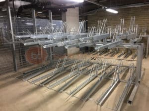 A metal bike rack structure with multiple upper and lower slots for holding bicycles is positioned indoors. The background shows industrial elements like metal fencing, pipes, and a concrete wall. The area appears to be a bike storage room or facility.