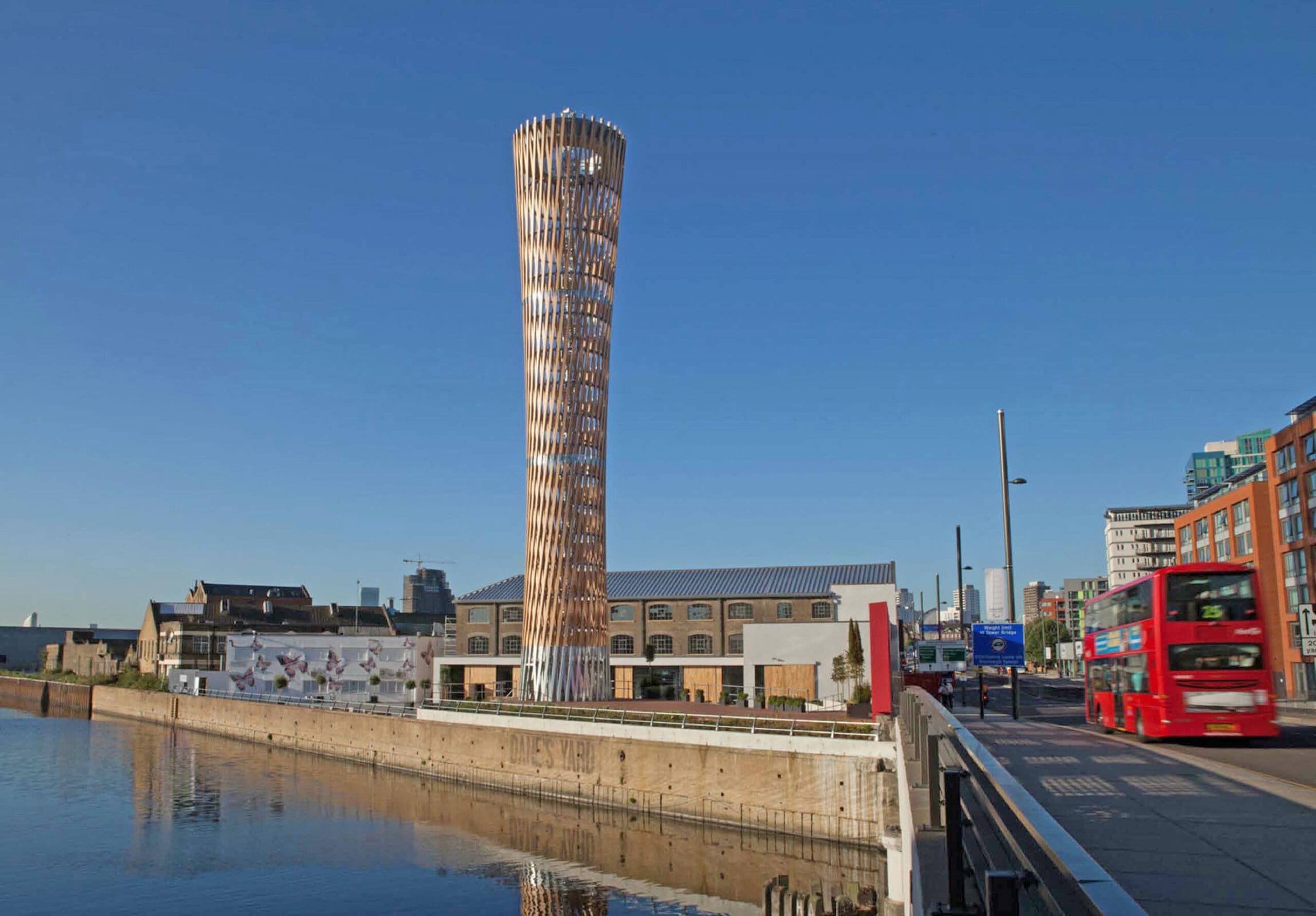 A tall, cylindrical, modern building stands by a waterfront under a clear blue sky. The structure features a unique design with a twisting facade. Nearby, a red double-decker bus is visible on the right, and several other buildings are in the background.