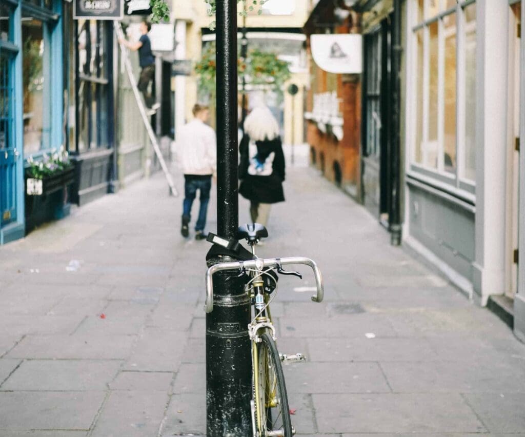 A narrow cobblestone street with a bicycle locked to a lamppost in the foreground. In the background, two people, a man and a woman, walk down the street while a worker stands on a ladder attending to a shopfront. The buildings are colorful and closely spaced.