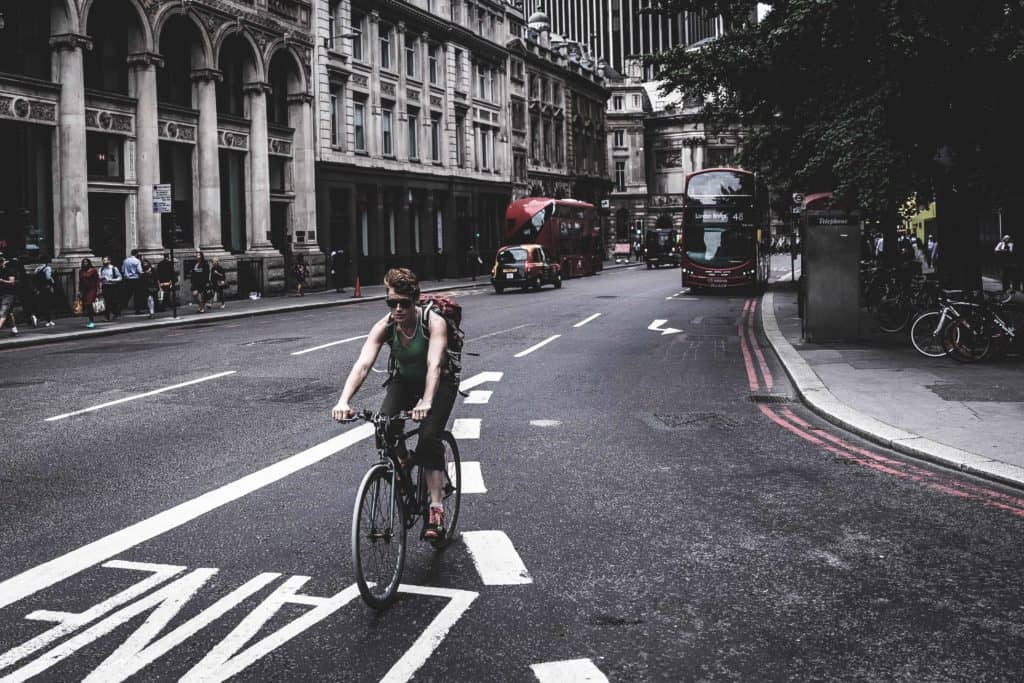 A cyclist riding in a designated bike lane on a busy city street with historic buildings and red double-decker buses in the background. Pedestrians are visible on the sidewalks, and there is a lush tree on the right side of the street.