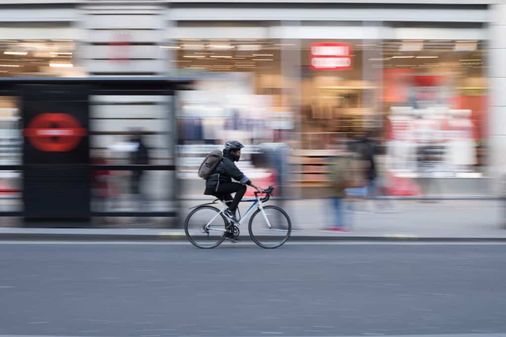 A cyclist in motion rides past a busy urban shopping area. The background is blurred, highlighting various storefronts and pedestrians, while the cyclist, wearing a helmet and backpack, is in sharp focus on the road.